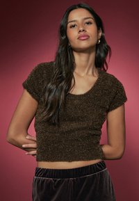 Brown, short-sleeve cropped sweater with a soft, fuzzy texture and shimmer, paired with dark, plush velvet pants against a red background.