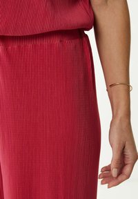Rode geplooide stof met een gestructureerd oppervlak, met een hoge taille en een zijnaad. Een dunne, gouden armband siert de pols.
