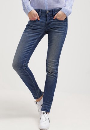 Personne portant un jean bleu ajusté, les mains dans les poches, avec des baskets blanches sur un fond clair uni.