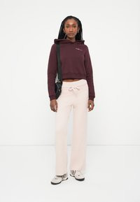 Bordový skrátený mikina s ružovým logom, kombinovaná so svetloružovými huňatými širokými nohavicami. Model má na sebe čiernu crossbody tašku a vzorované tenisiky.