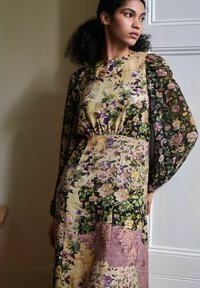Kukkaistettu mekko, jossa on värien sekoitus: vihreä, violetti ja kultainen. Pitkät hihat ja istuva vyötärö, jossa on teksturoitu kangas.