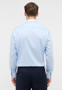 Camicia a maniche lunghe azzurro chiaro con colletto standard, texture liscia, polsini a due bottoni e dettaglio della spalla sul retro. Indossata con pantaloni scuri.