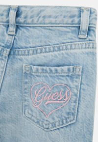 Svetlo modri jeans iz denima imajo zadnji žep z rožnato vezenino v obliki srca z napisom "Guess." Tkanina ima zbledel, teksturiran videz.