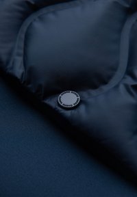 Close-up af mørkt polstret stof med en rund knap mærket "OMBRE" flere gange, der fremhæver tekstur og syningsdetaljer.