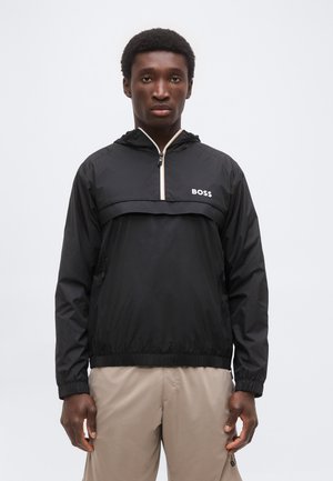 Noor mees kannab musta BOSS tuulejopet lukuga krael ja beežeid pükse, seisab ühevärvilise heledahall tausta ees.