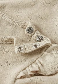 Tissu tricoté beige présentant un motif texturé. Comprend des boutons-pression argentés et un bord froncé, mettant en avant un matériau doux et léger.