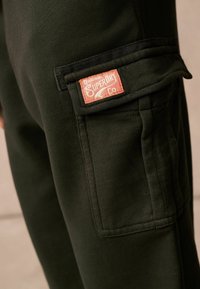 Black cargo pants featuring an embroidered orange label, front pocket, and a soft, textured fabric. Focus on utility and comfort.