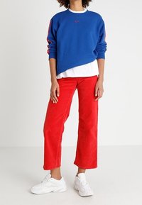 Blå crewneck-tröja med röd Nike-logotyp och detaljer, tillsammans med röda kordfløjlsbyxor med vida ben och vita sneakers.