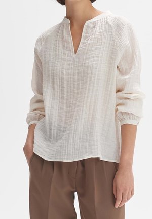 Frau trägt eine lange, lockere, weiße Bluse mit Textur, einem V-Ausschnitt und geknöpften Ärmeln, kombiniert mit brauner Hose.