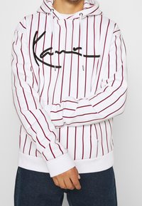 Felpa con cappuccio bianca con strisce verticali rosse e logo firma nero. Include una tasca frontale e cappuccio con coulisse. Polsini elasticizzati.