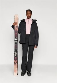 Naine mustas suusajakis ja pükstes hoiab vertikaalselt paari suuski, tal on jalas mustad suusasaapad ja roosa aluskiht.