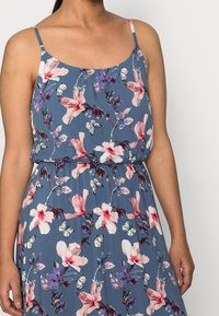 Kvinna i en blå ärmlös klänning med mönster av rosa och lila blommor och fjärilar, med tunna axelband och en rynkad midja.