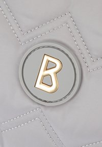 Šedá prošívaná látka s prošitým cikcak vzorem a kulatým logem se zlatými akcenty zobrazujícím stylizované písmeno 'B'.