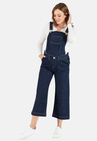 Dunkelblaue Denim-Latzhosen mit weitem Bein, verstellbaren Trägern, einer Vordertasche und goldenen Hardware-Details, getragen über einem weißen Langarmshirt. Weiße Turnschuhe.
