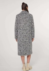 Langer grauer Mantel mit einer flauschigen Textur, mit einem Reverskragen und geradem Schnitt. Er hat ein mehrfarbiges, gepunktetes Muster und mittellange Ärmel.