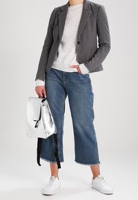 Blazer gris à fines rayures porté sur un pull léger, associé à un jean bleu raccourci et des baskets blanches. Un sac à dos argenté métallique complète la tenue.