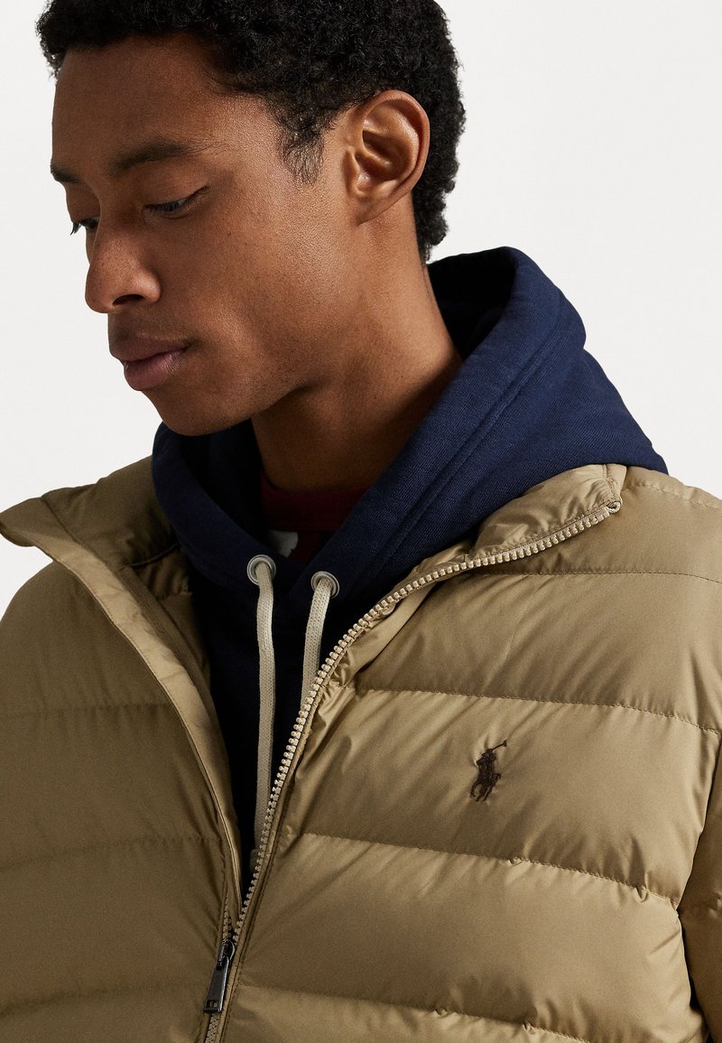 Beige donsjacket met een horizontaal gestikt ontwerp, een ritssluiting aan de voorkant en een marineblauwe hoodie met aansnoerkoorden eronder. Voorzien van een logo-embleem.