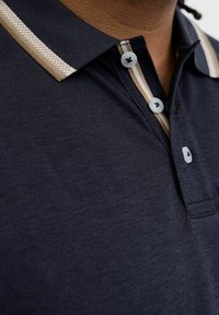 Tuvie tuvumā redzams tumši zils polo krekls ar smilškrāsas un baltām svītrām apkaklē un dekoltē daļā, kurā ir redzami trīs baltie pogas.