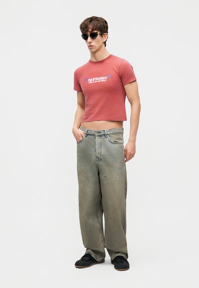 Young man wearing black sunglasses, red cropped T-shirt reading "I'M STRAIGHT (this is an old shirt)," wide faded jeans, and black shoes.