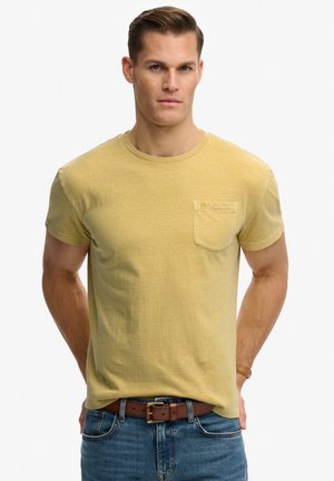 Hombre vestido con camiseta de manga corta color mostaza con bolsillo, vaqueros azules y cinturón de cuero marrón, de pie contra un fondo blanco.