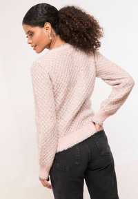 Világos rózsaszín kötött pulóver, textúrált gyémánt mintázattal, bordás mandzsettákkal és szegéllyel. Sötét magas derekú nadrággal párosítva.