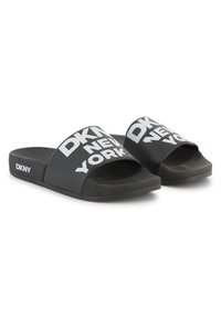 Chanclas negras con una tira ancha que presenta el texto blanco "DKNY NEW YORK". La plantilla y la suela exterior texturizadas también son negras y planas.