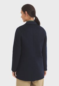 Abrigo de lana en color marino con un cuello estructurado, mangas largas y corte recto. Presenta una costura en la parte trasera central y un diseño limpio y minimalista.
