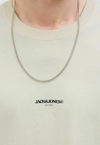 Béžové tričko s krátkým rukávem a kulatým výstřihem, na kterém je černé logo "JACK & JONES" a stříbrný řetízek na pozadí hladké textury látky.