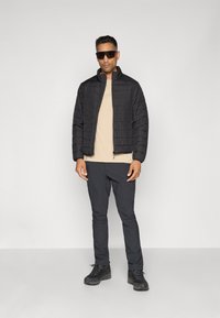 Doudoune noire avec fermeture éclair à l'avant et motif matelassé, associée à un t-shirt beige et à un pantalon noir ajusté. Bottes de randonnée noires.
