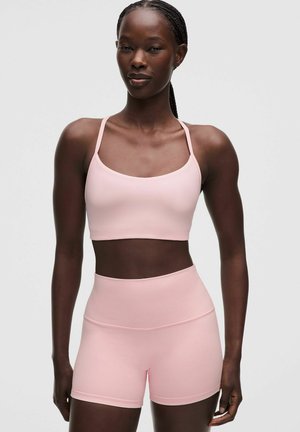 Donna con reggiseno sportivo rosa chiaro e pantaloncini abbinati a vita alta, in piedi davanti a uno sfondo grigio semplice.