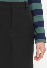Jupe crayon noire en tissu texturé avec une finition lisse, dotée d'un pli avant et d'une poche latérale, associée à une chemise rayée verte et marine.