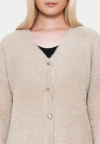 Beige, bolyhos kardigán V-nyakkal, négy ezüst gombbal az elején és puha textúrával. Fekete blúz felett viselve.