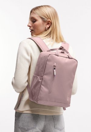 Zaino rosa chiaro a forma rettangolare, realizzato in tessuto liscio, presenta una tasca anteriore con zip e una tasca laterale, spallacci regolabili e un manico ad anello.