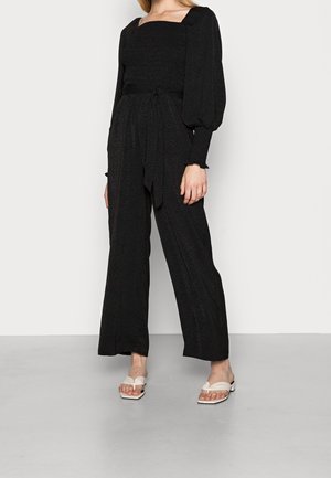 Frau trägt einen schwarzen Jumpsuit mit langen Ärmeln, Puffärmeln, Bindegürtel und weiten Beinen sowie weiße Sandalen mit Absatz, steht auf einem grauen Boden.