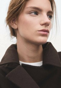 Bruine wollen jas met een brede kraag, gedragen over een wit kledingstuk. Het onderwerp heeft een lichte huid en eenvoudige oorbellen. Natuurlijke haarttextuur, neutrale achtergrond.