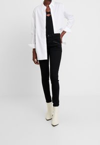 Bílá košile s knoflíky přes černý top, spárovaná s černými skinny džíny a krémovými kotníkovými botami se zúženým podpatkem. Hladké tkaniny a čisté linie.