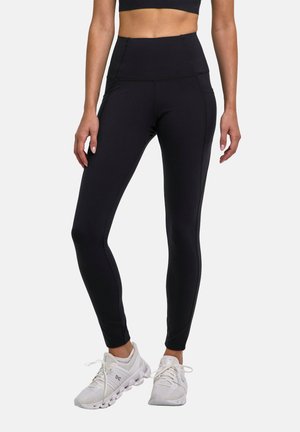 Femme portant des leggings noirs taille haute et des chaussures de course blanches, debout sur un fond uni.