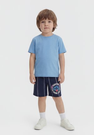 Fiatal fiú világosbarna hajjal, világoskék pólóban, sötétkék rövidnadrágban, amelyen rajzfilmfigura van, fehér zokniban és fehér sportcipőben.