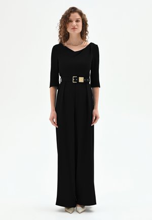 Femme aux cheveux bouclés portant une combinaison noire à manches trois-quarts, une ceinture large avec une boucle dorée et des chaussures argentées pointues.