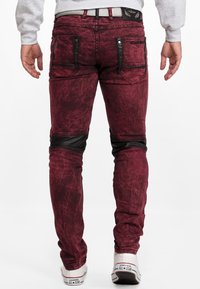 Jean slim rouge texturé avec poches zippées noires et accents aux genoux, associé à des baskets blanches, montrant une vue de dos.