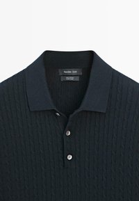 Tmavě modré polo tričko z 100% bavlny s texturovaným vzorem copánkového pletení, dvojitým knoflíkovým límečkem a žebrovaným límcem.