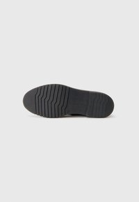 Suela de zapato de vestir negro con un patrón de tracción en zigzag y el nombre de la marca "bugatti" grabado en un fondo blanco liso.