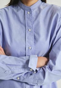 Chemise boutonnée légère en violet, avec un col chinois, des manches longues et des boutons blancs. Le tissu semble lisse avec un minimum de brillance et une coupe décontractée.