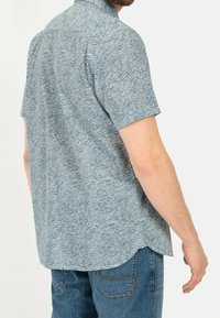 Camisa de manga corta azul con estampado de olas, hecha de tejido ligero, con un cuello clásico y un dobladillo curvado en la parte trasera.