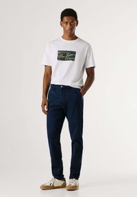 White t-shirt with blue logo, navy trousers, and beige sneakers. T-shirt features a rectangular graphic design; trousers have a sleek fit.
