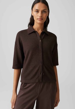 Frau trägt ein dunkelbraunes, kurzärmliges Strickhemd mit Knöpfen und passende Hose, steht vor einem schlichten hellgrauen Hintergrund.