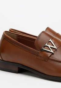 Bruine leren instappers met een lage hak, voorzien van een zilverkleurig "W" logo accent. Gladde textuur met stikdetails langs de bovenkant.