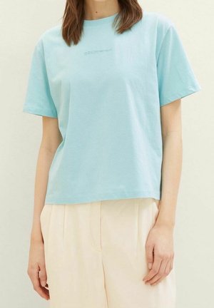 Persoon die een lichtblauw T-shirt met korte mouwen en losse, hooggetailleerde crèmekleurige broek draagt, staand tegen een effen achtergrond.