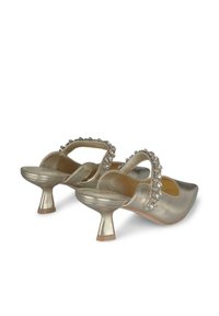 Chaussures slip-on dorées métalliques avec un bout arrondi, un petit talon et une sangle ornée de pierres précieuses transparentes sur le devant.