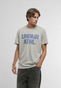 Šedé bavlněné tričko s výrazným modr�ým nápisem "UNFAIR ATHL." Krátké rukávy, kulatý výstřih, volný střih, hladký materiál.